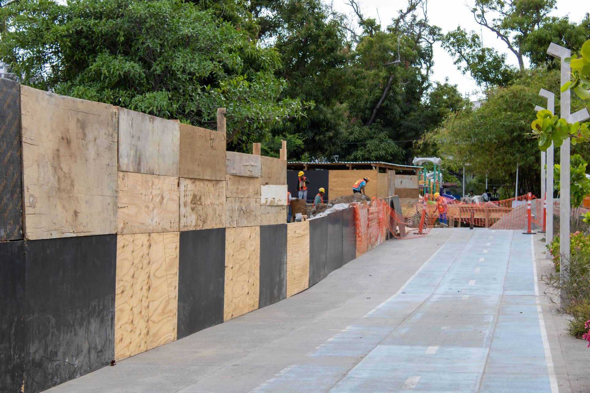Outdoor walkway beside a patchwork wooden wall, with orange fencing and trees in the background.
