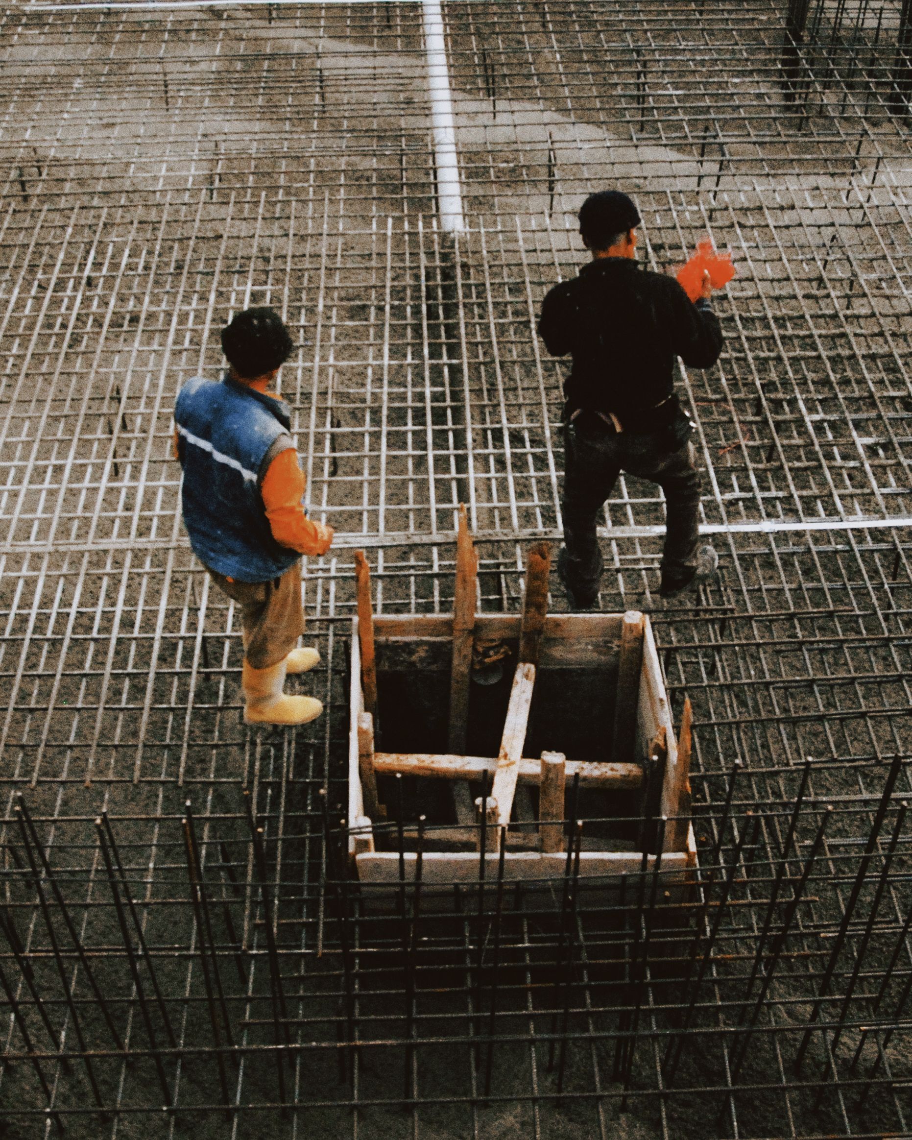 Two construction workers stand on a rebar grid beside a concrete form on a building site.