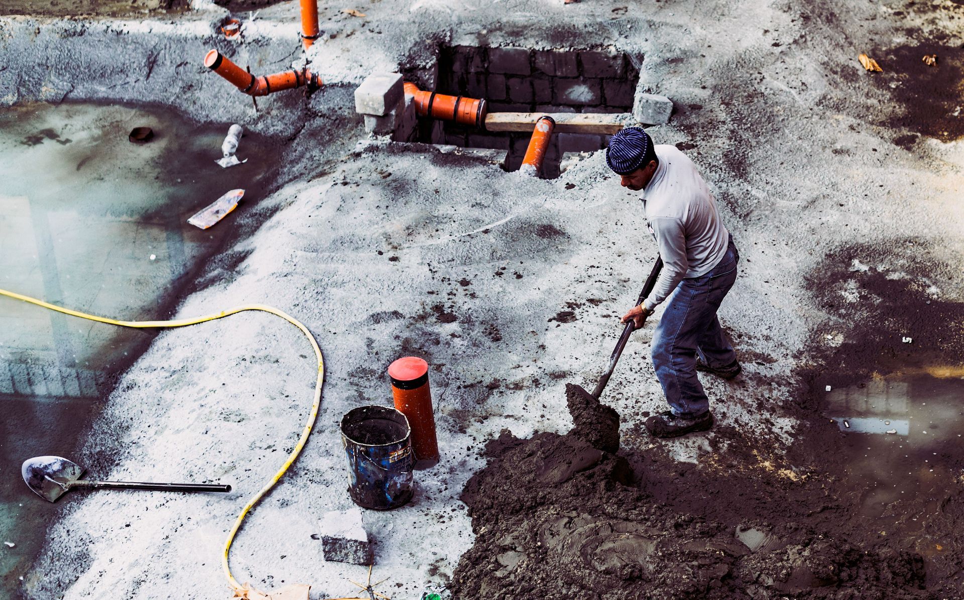 Worker shoveling concrete at a construction site with pipes and wet ground nearby