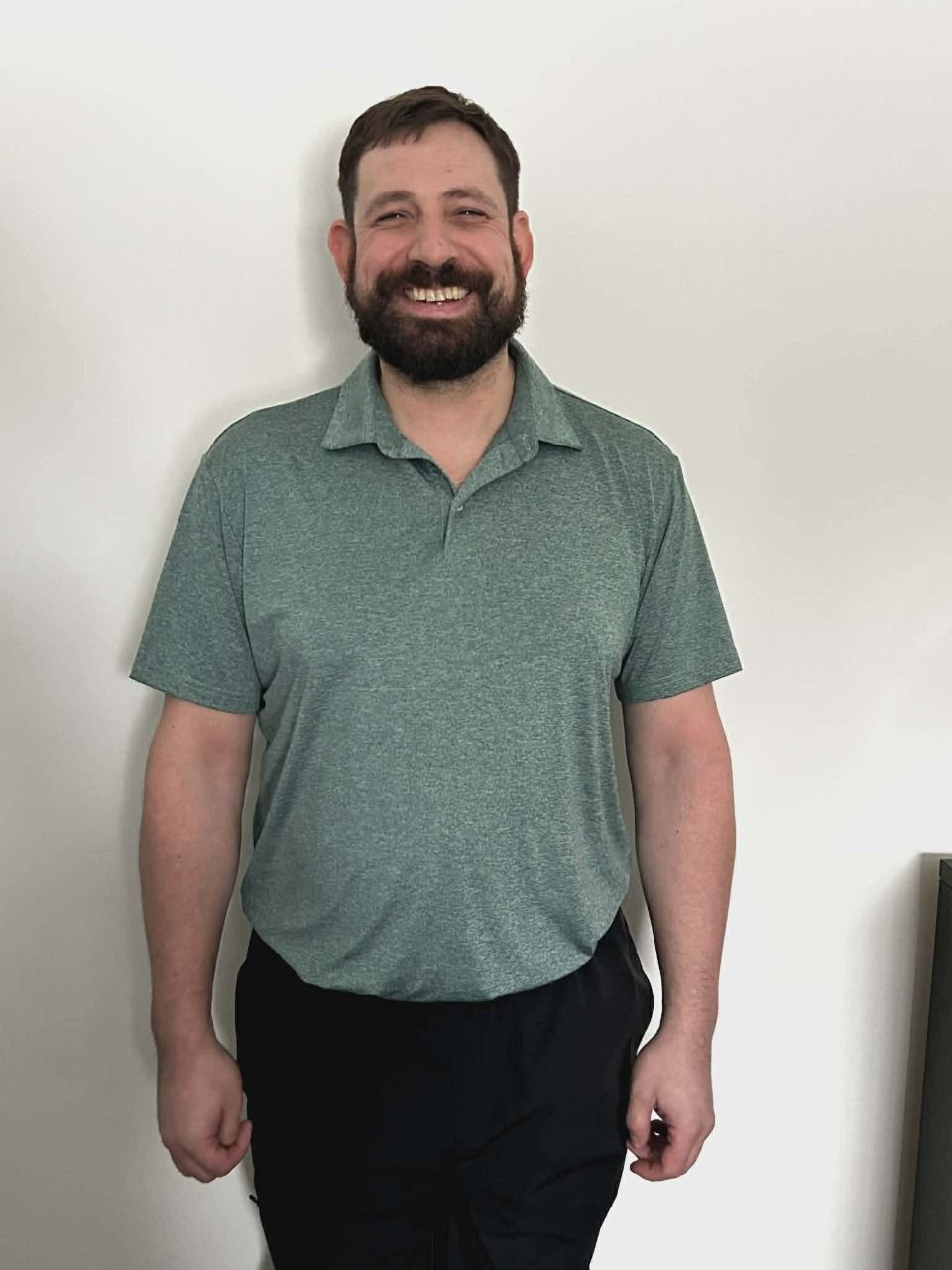 Smiling man standing against a white wall, wearing a green polo shirt and black pants.