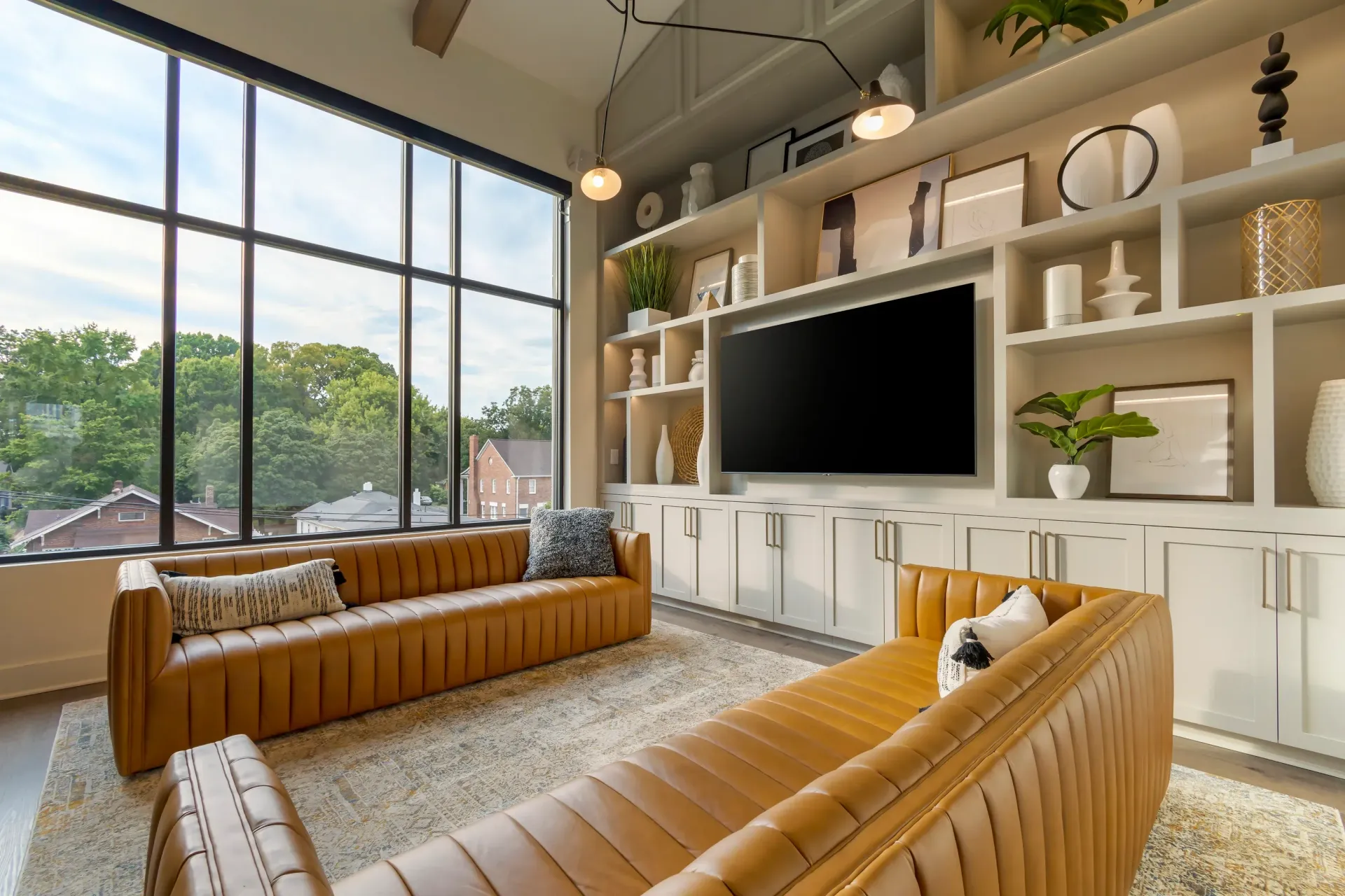 Bright apartment living room with large windows, tan leather sofas, and built-in white shelving around a TV.