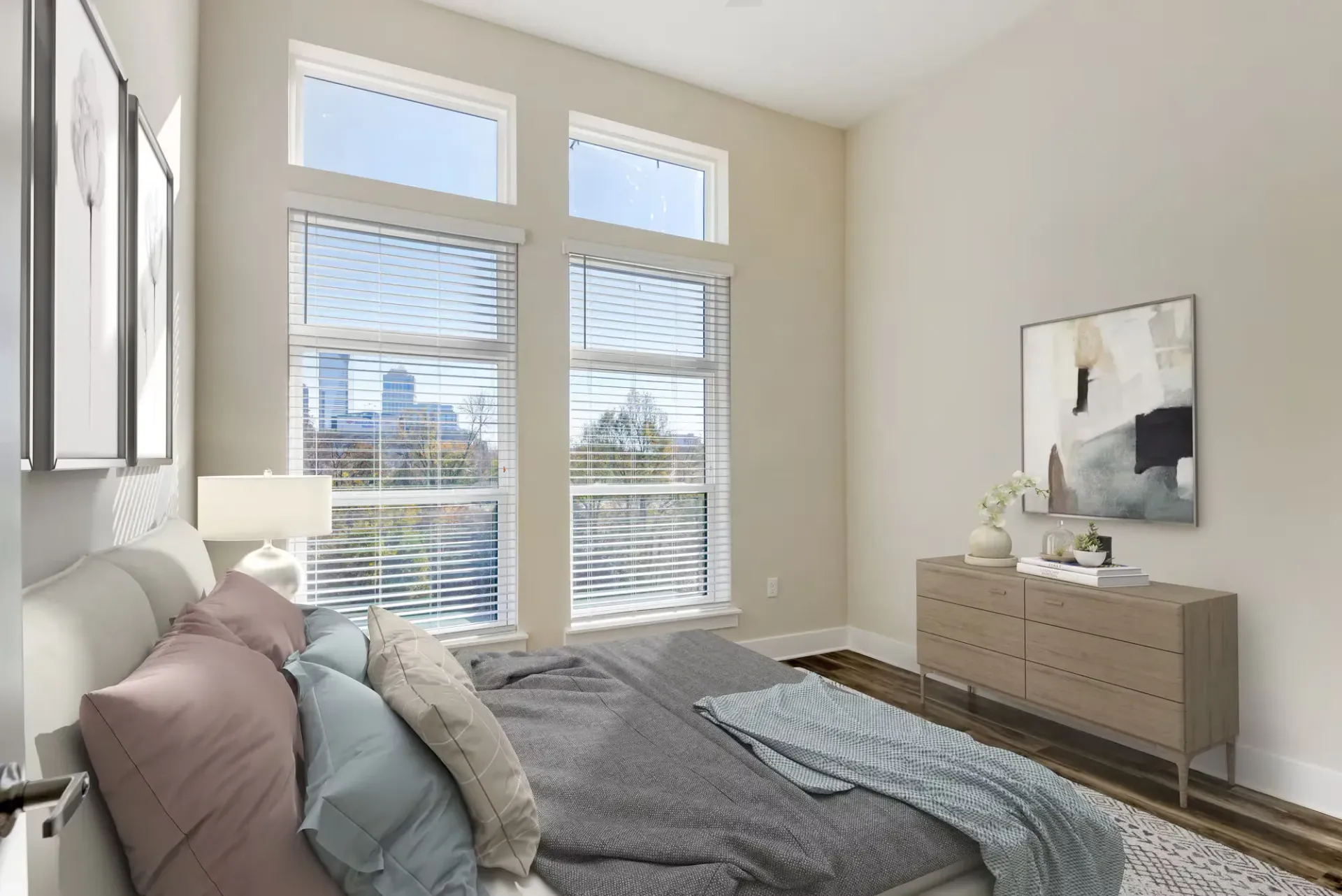 Bright bedroom with tall windows, blinds, a bed with pastel pillows, and a wooden dresser.