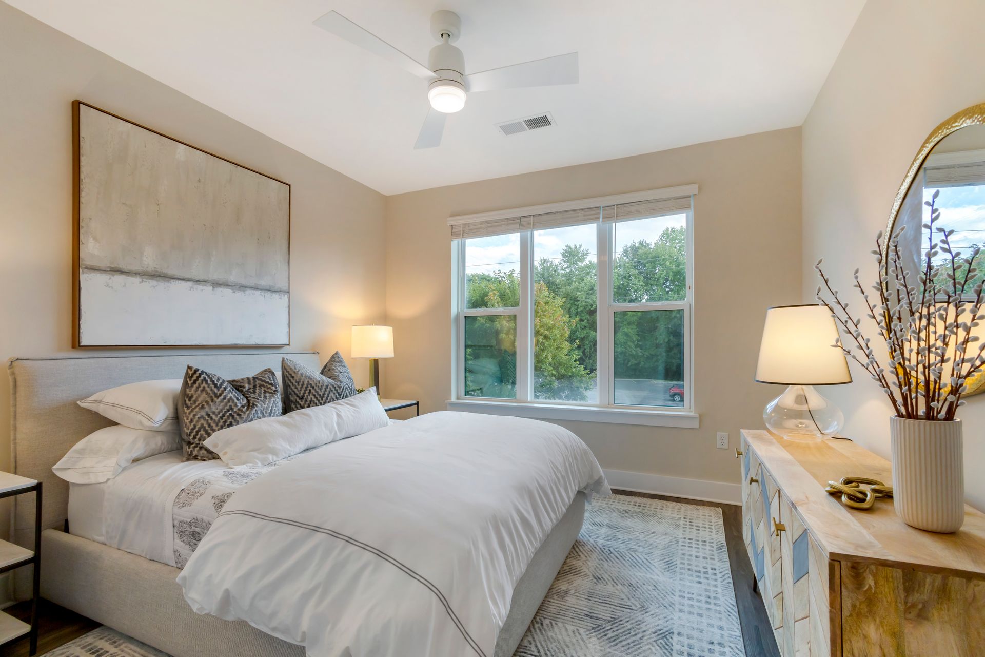 Bedroom with a bed, dresser, large window, and abstract art. Neutral tones, well-lit.
