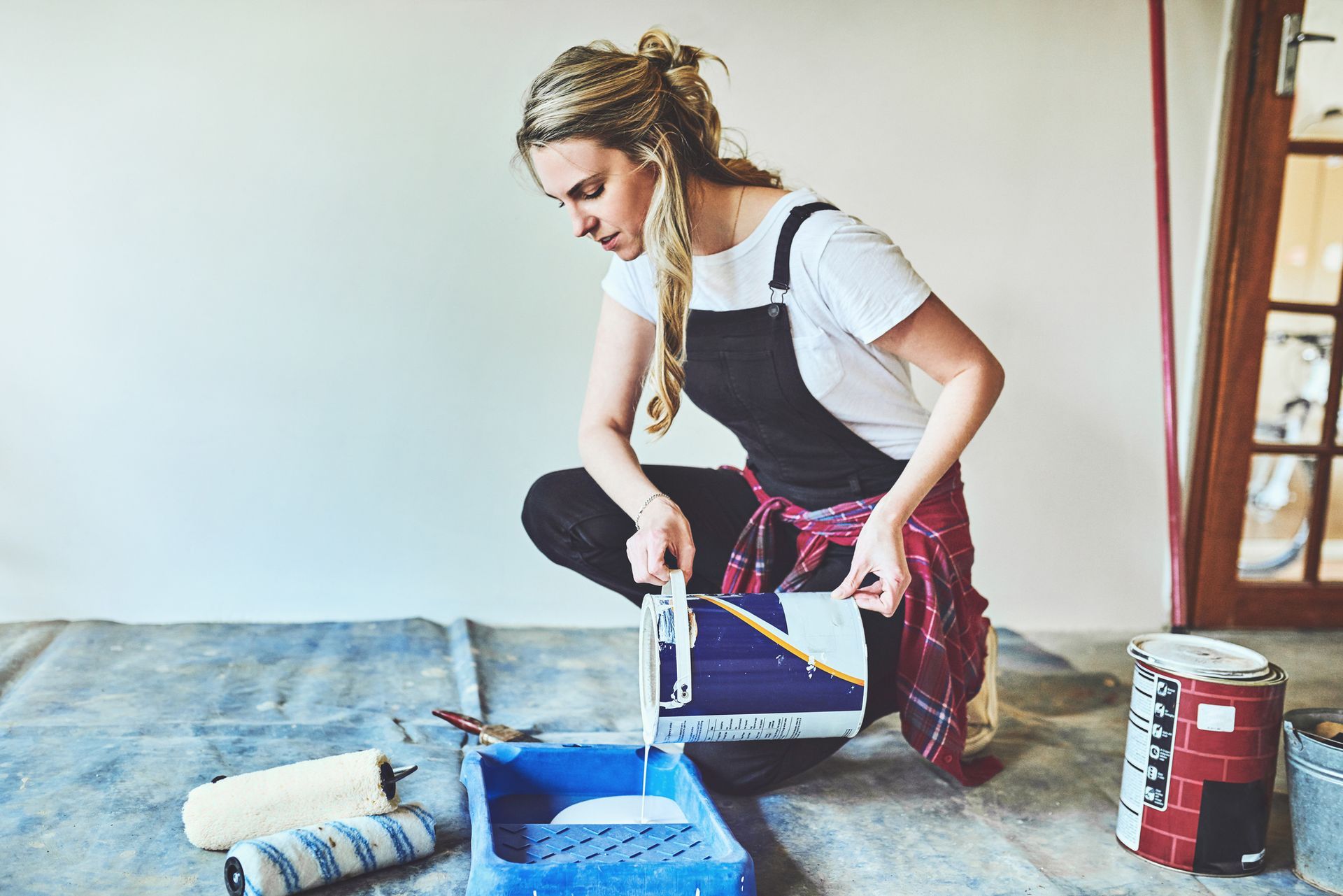 Woman pouring paint into a paint tray. She's wearing overalls and is in a room.
