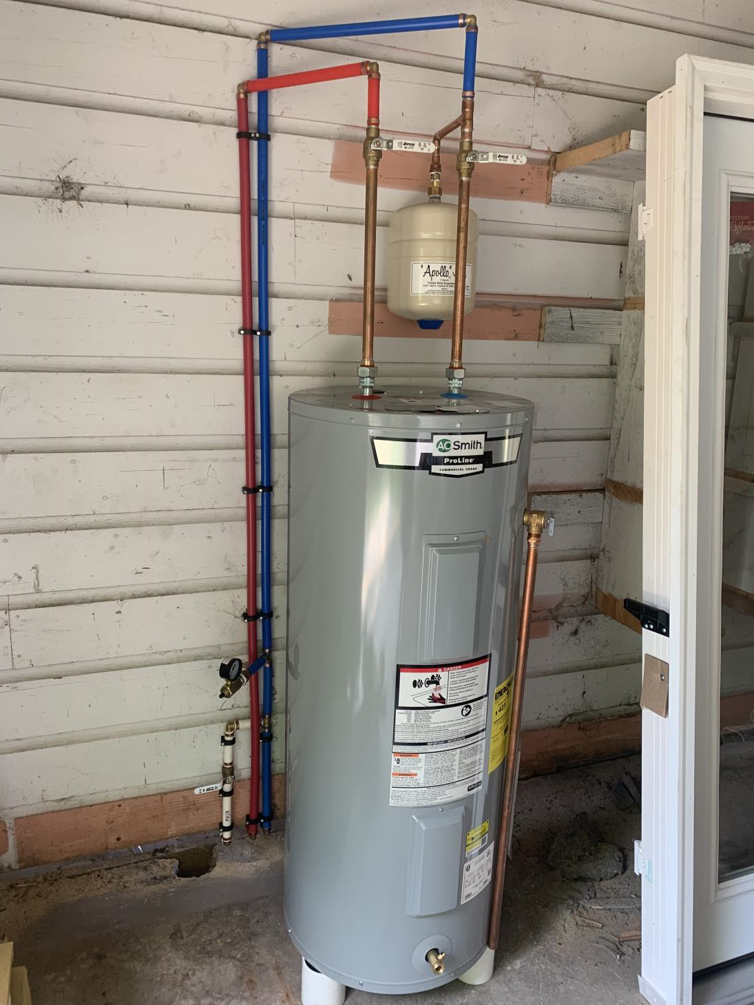 Water heater with copper pipes, expansion tank, and blue/red PEX plumbing on a weathered wall.