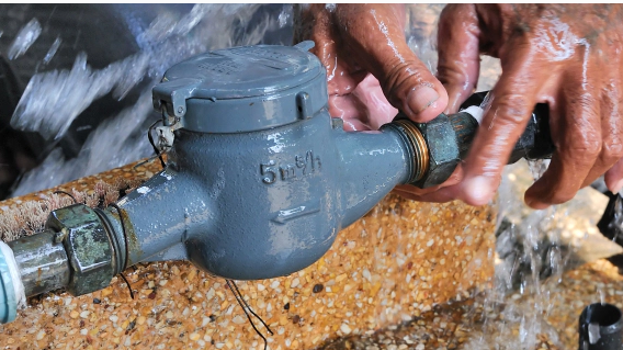 Hands tightening a fitting on a leaking water meter; water sprays outward.