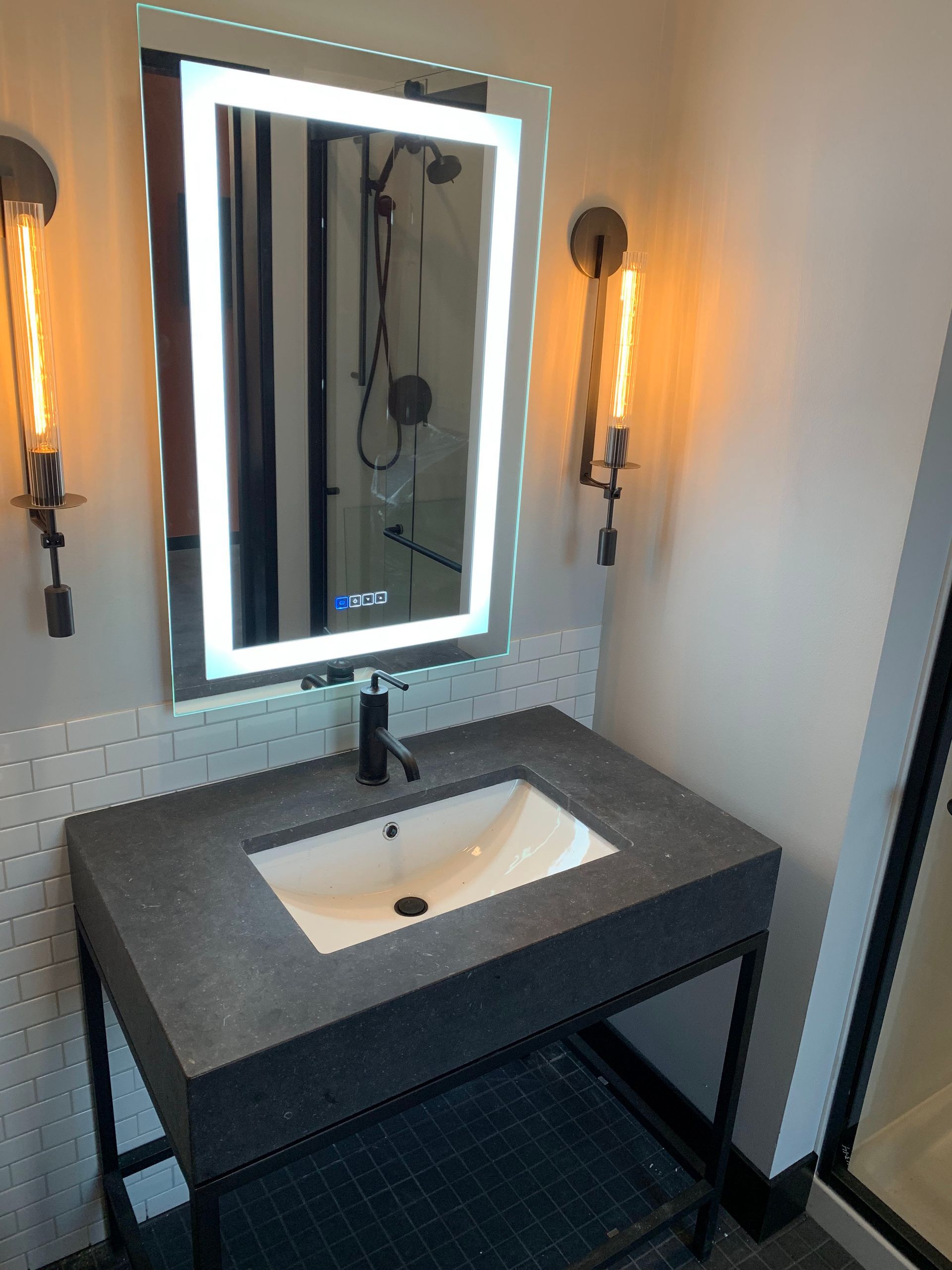 Bathroom with lit mirror, dark countertop, and industrial sconces.