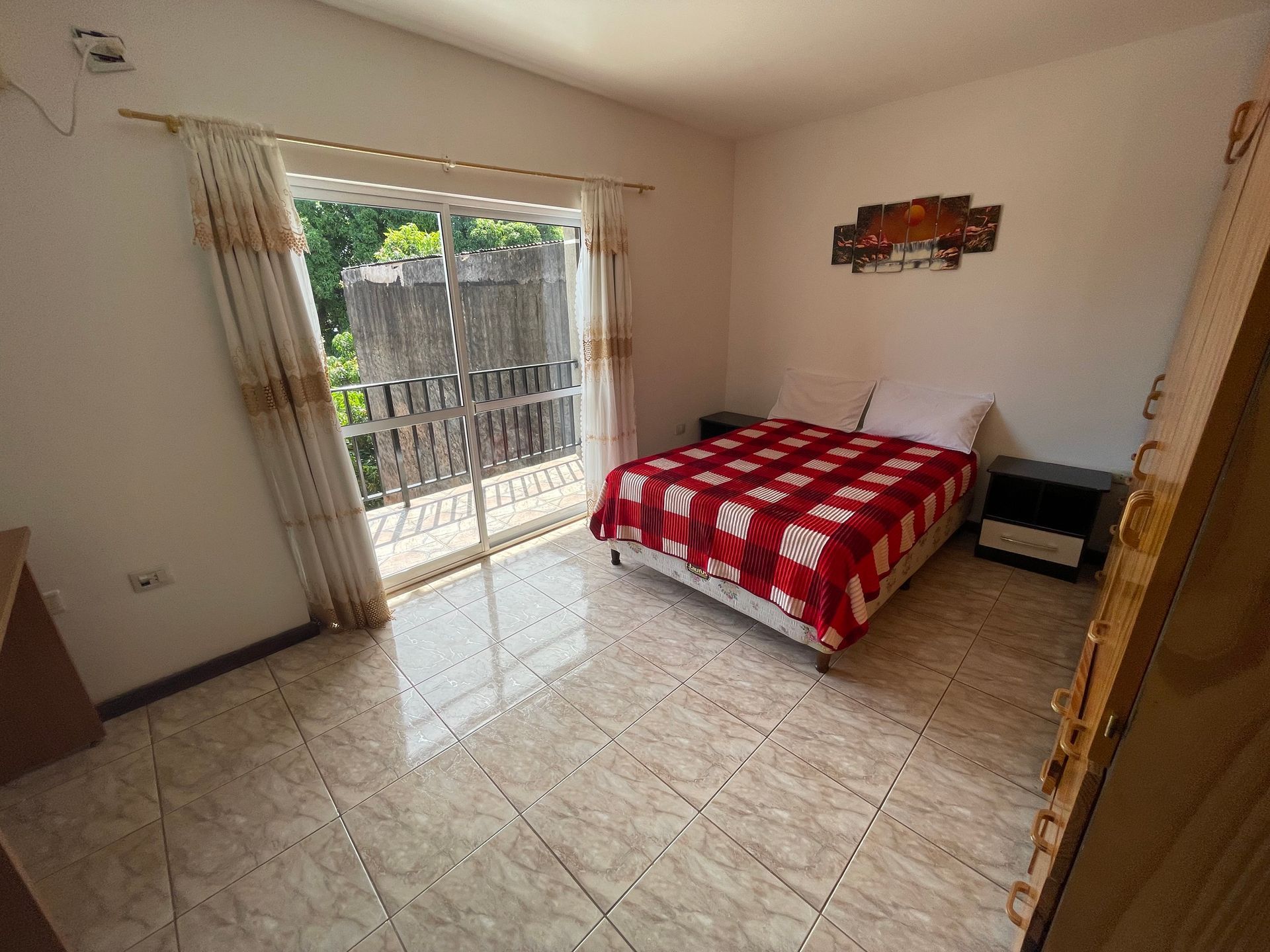 Bright bedroom with a red bedspread, tiled floor, and sliding glass door to a balcony