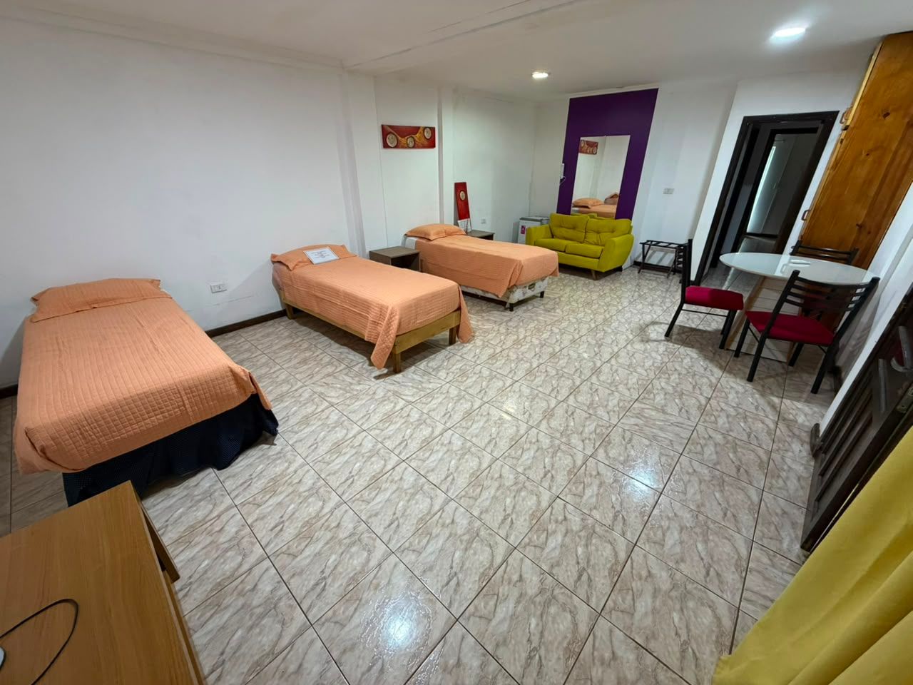 Shared dorm room with three single beds, tiled floor, desk, chairs, and a yellow dresser.