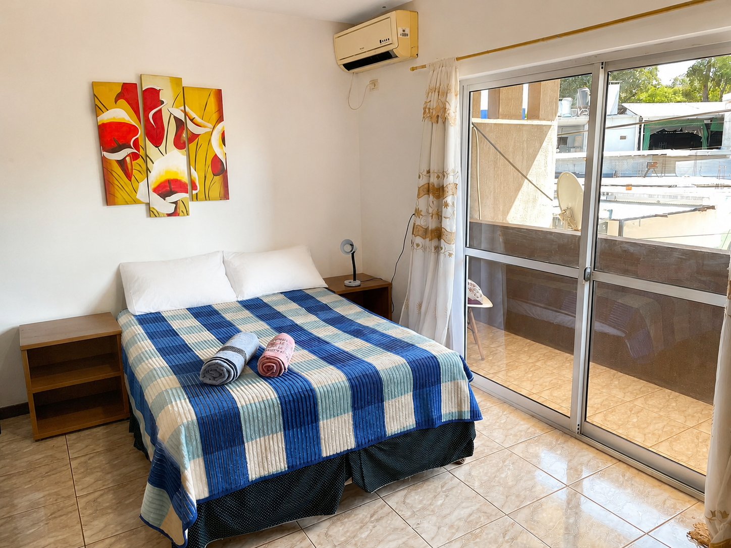 Small bedroom with blue-checkered bedspread, bedside tables, wall art, and sliding glass doors to a patio.