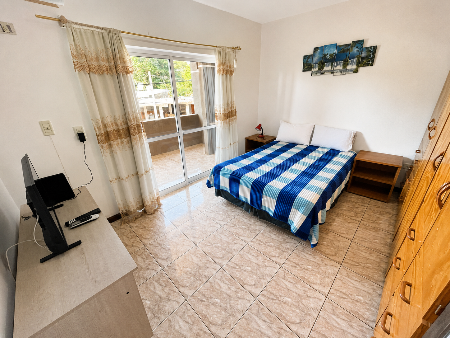 Bright bedroom with a blue checkered bedspread, sliding glass doors, TV, and wooden dresser.
