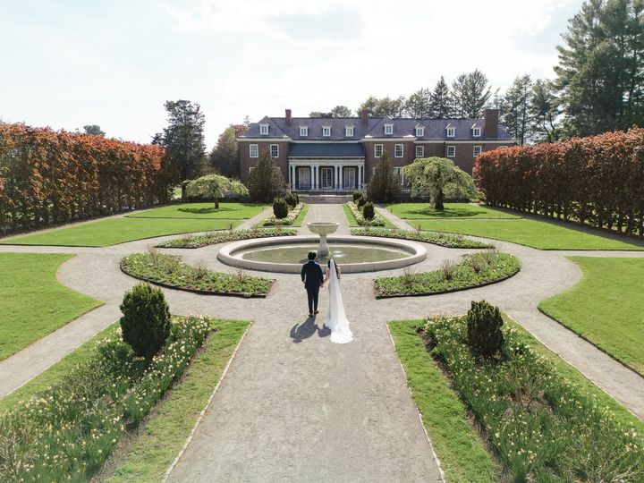 A bride and groom are walking down a path in front of a large luxury house.