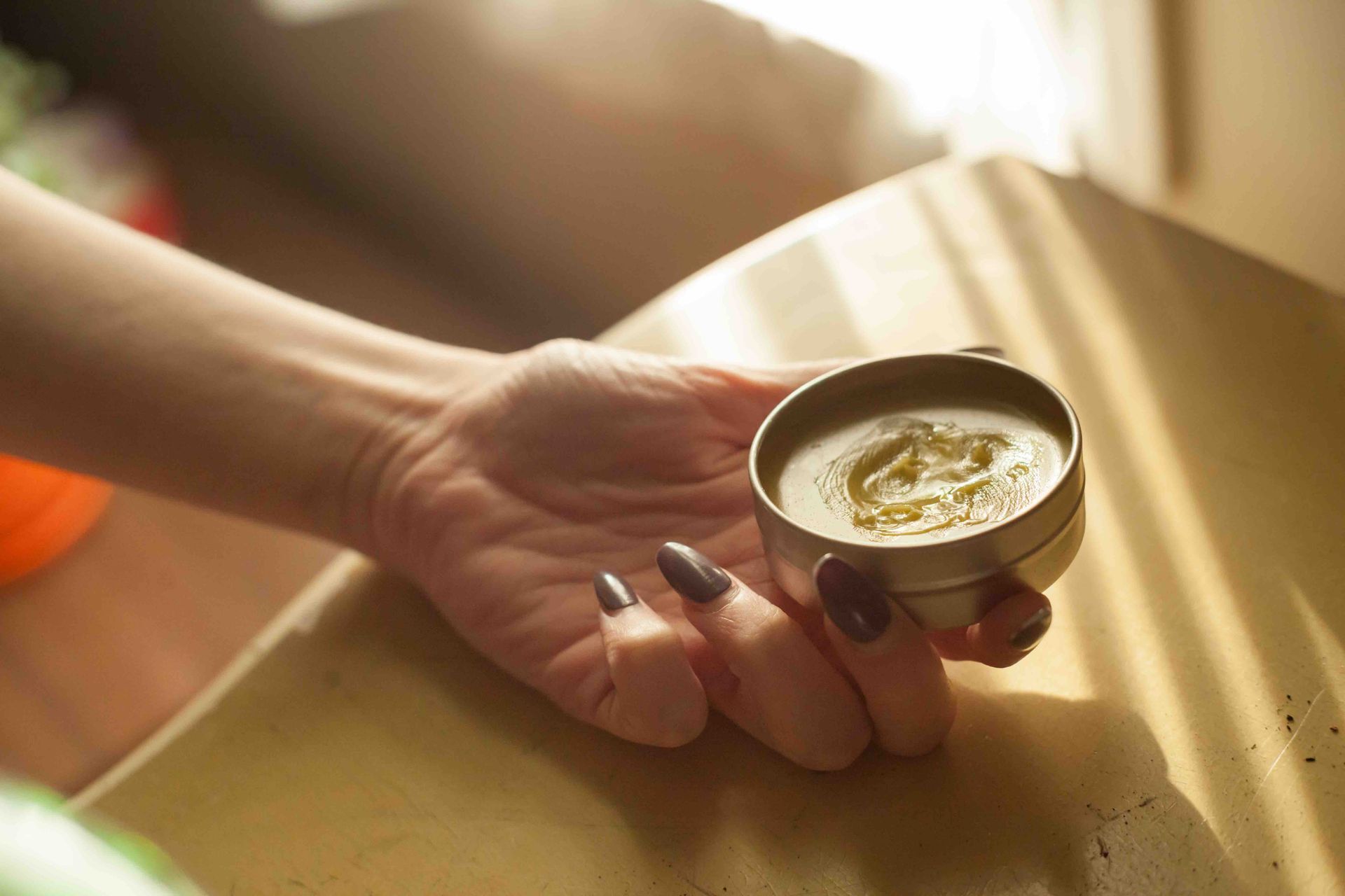Hand holding a small tin of cream on a sunlit wooden table
