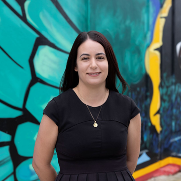 Smiling woman with dark wavy hair in a black top, seated indoors near a bright window Woman standing before a colorful mural with teal, black, and yellow geometric shapes.