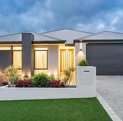 Modern single-story house with lit windows, dark garage, white fence, and trimmed lawn at dusk Modern single-story house with lit windows, dark garage, white fence, and trimmed lawn at dusk