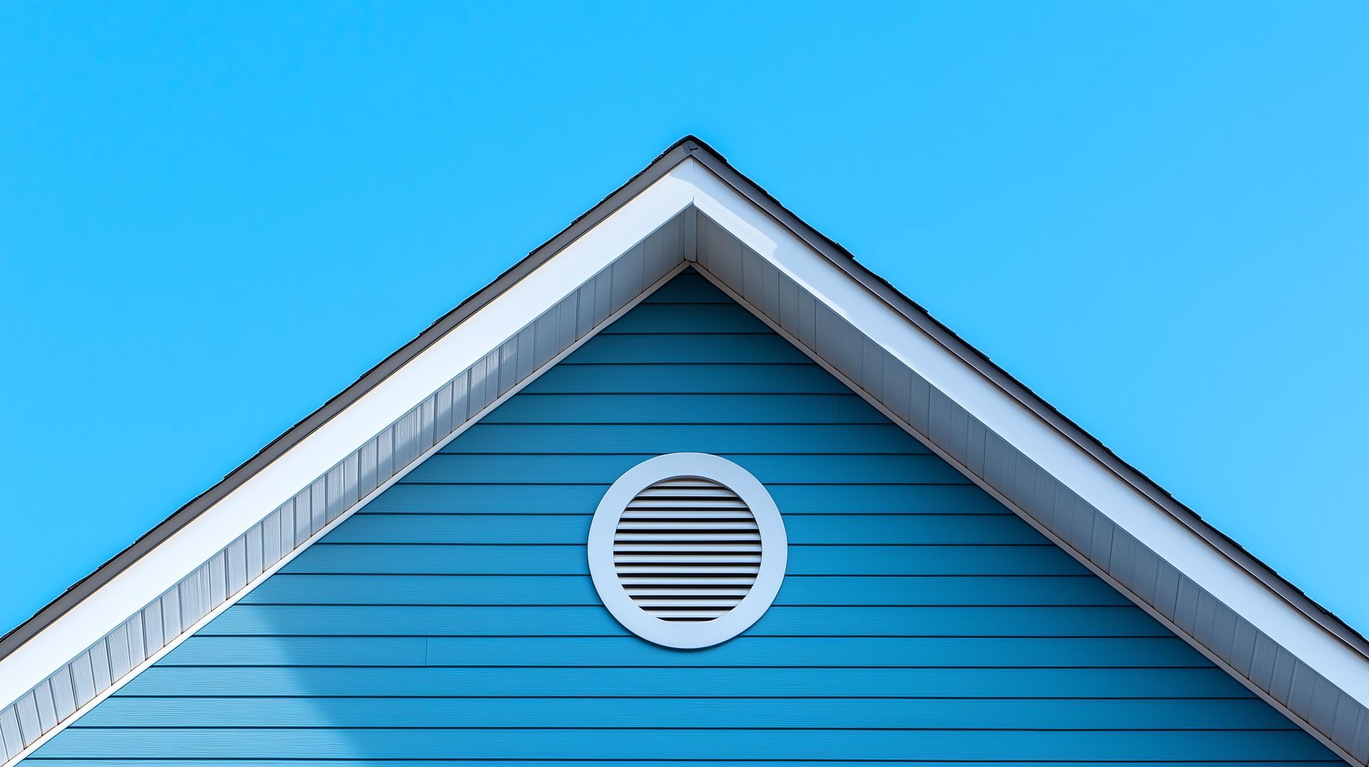 Blue house gable with white trim and round vent against a clear sky