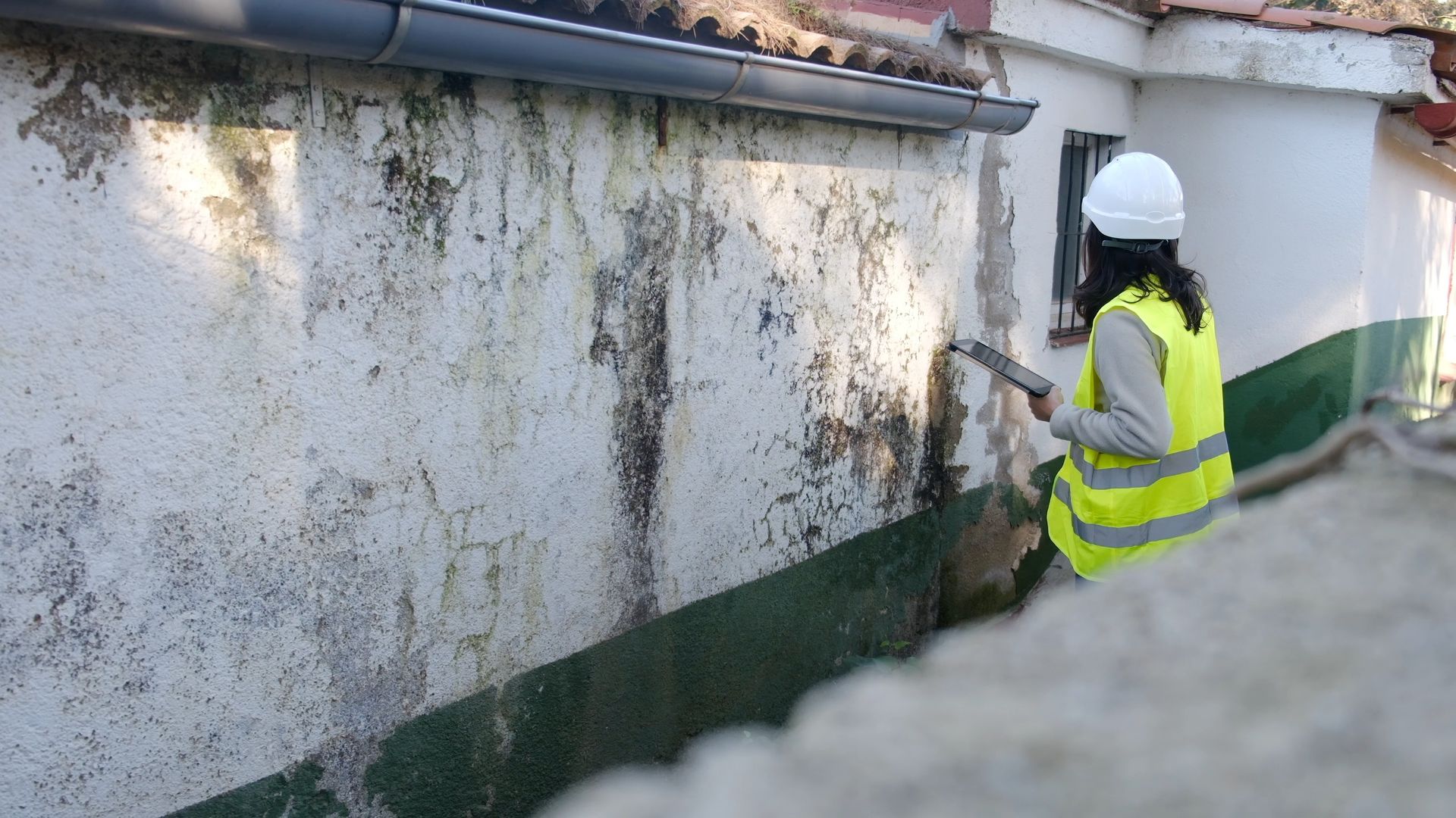 Worker in a white hard hat and yellow vest inspecting a stained exterior wall with a hose.