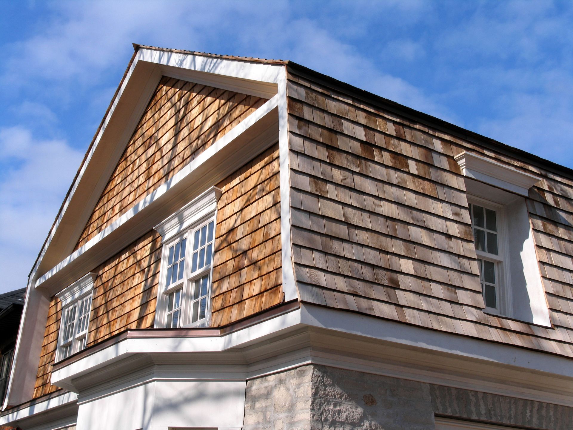 Corner of a house with tan shingle siding, white trim, and windows against a blue sky