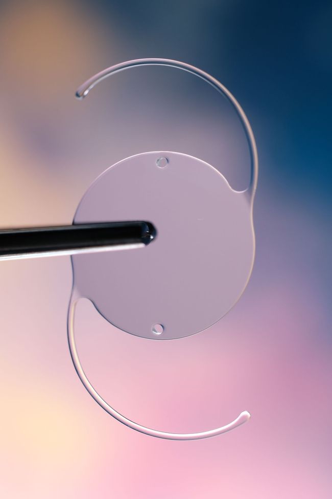 Lente intraocular sostenida con pinza; lente circular transparente con soportes curvos, sobre fondo colorido.