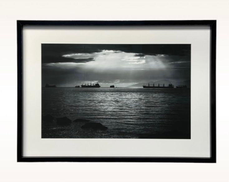A framed black-and-white photograph shows cargo ships silhouetted on a calm sea under a dramatic, light-streaked sky.