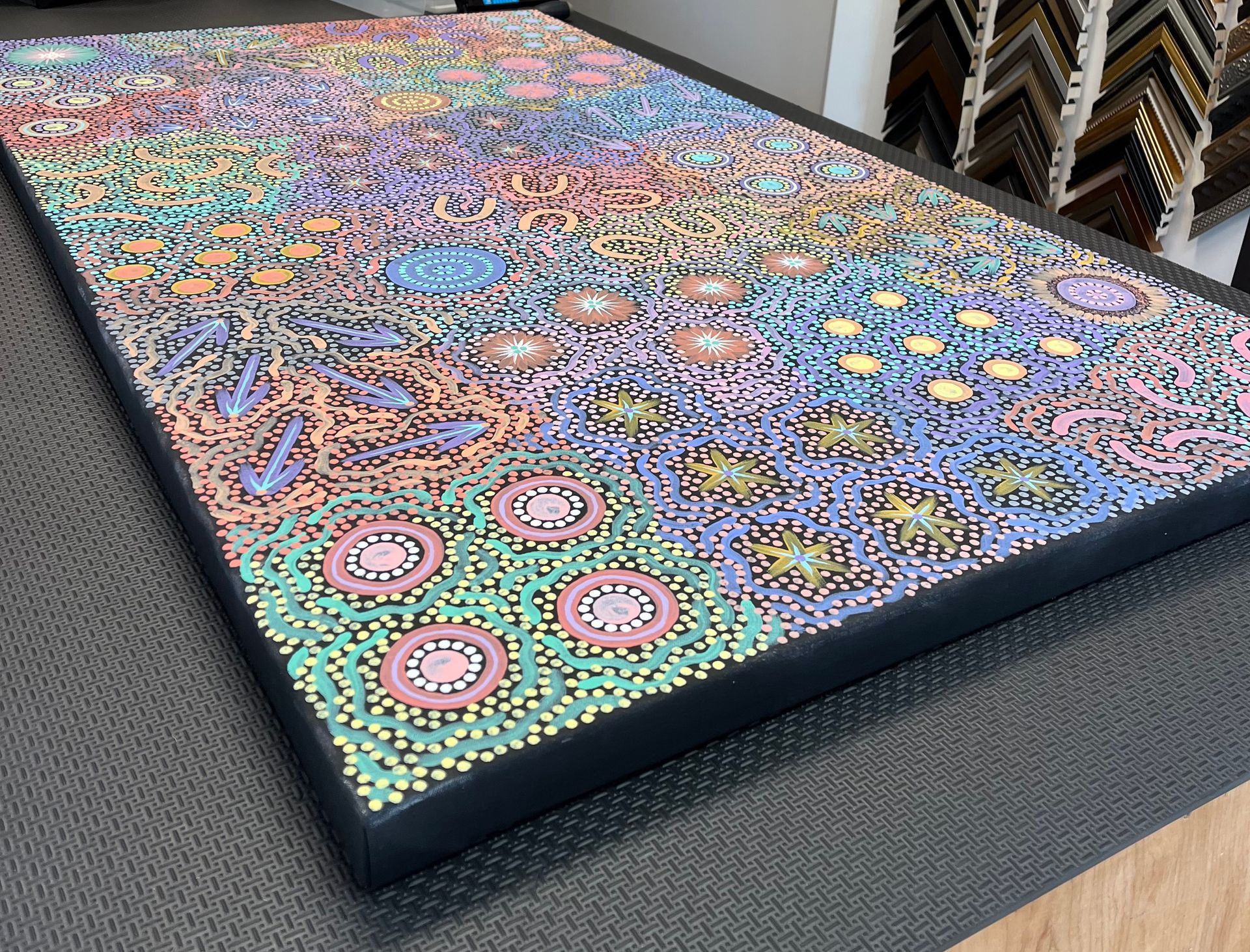 A rectangular canvas featuring a dense, colorful Aboriginal dot painting, set on a textured surface in a frame shop.