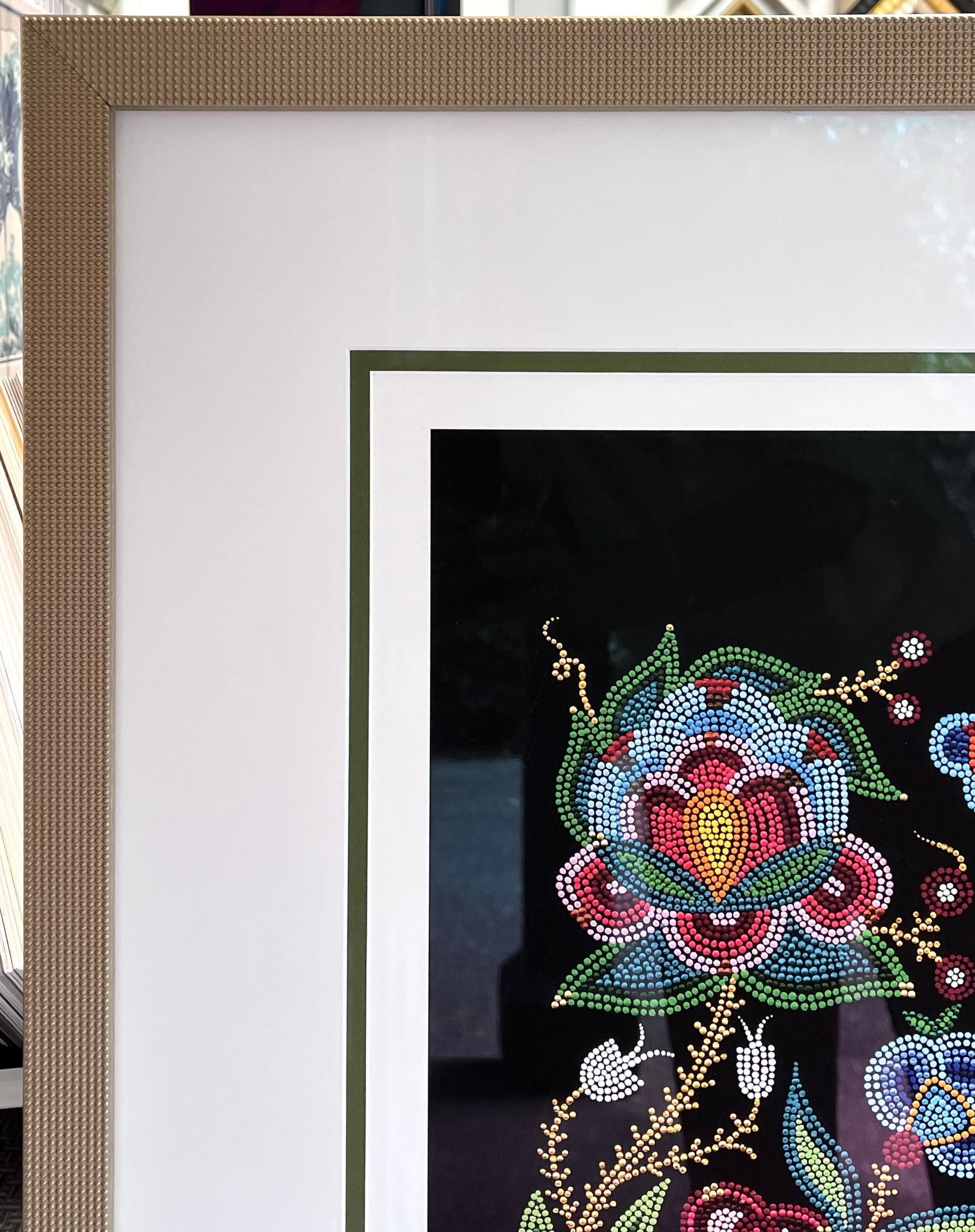 A framed, multi-colored floral embroidery on a black background, with a white matte and a textured gold-toned frame.