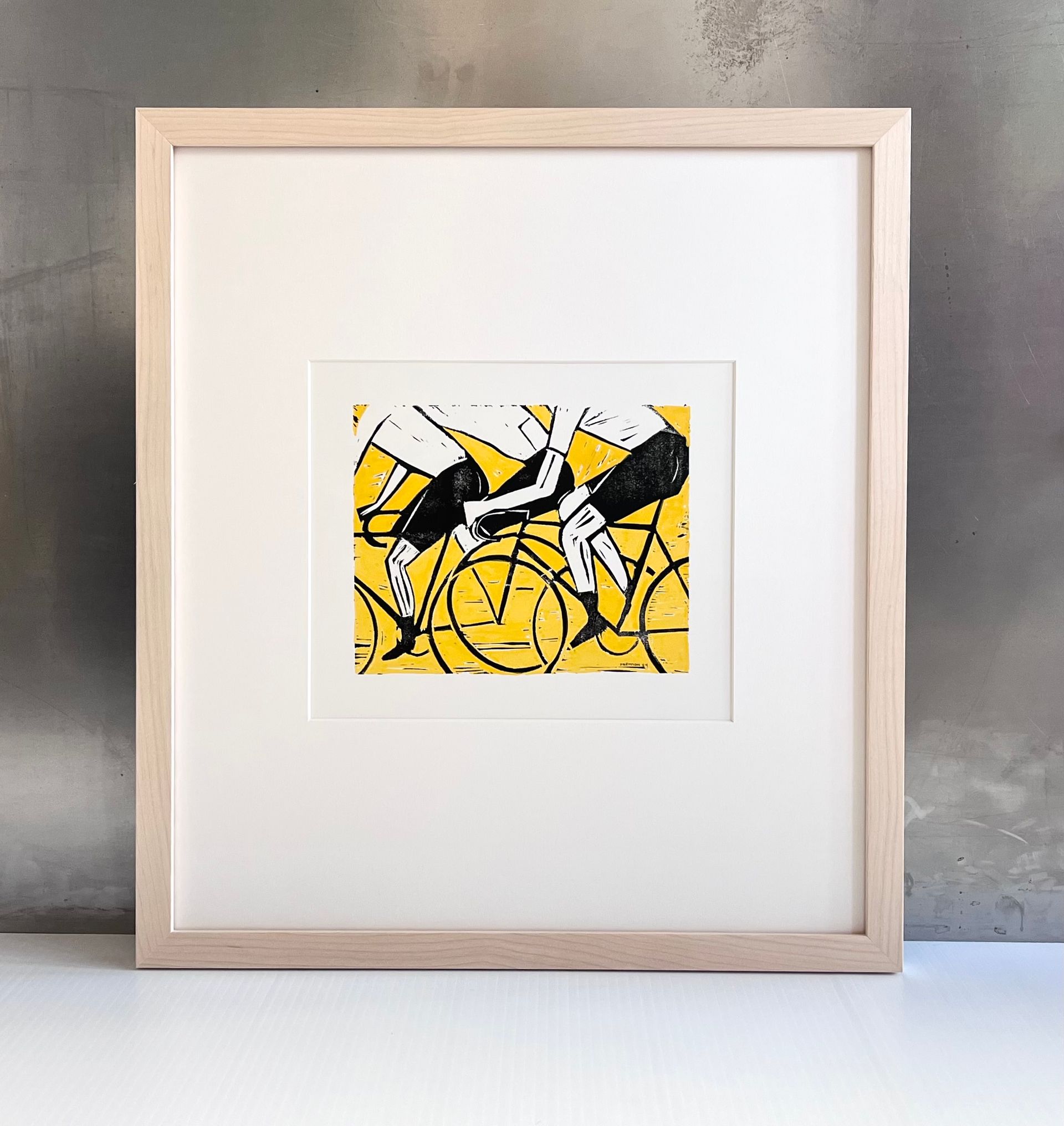 A framed linocut print featuring two cyclists in black on a yellow background, displayed against a gray wall.