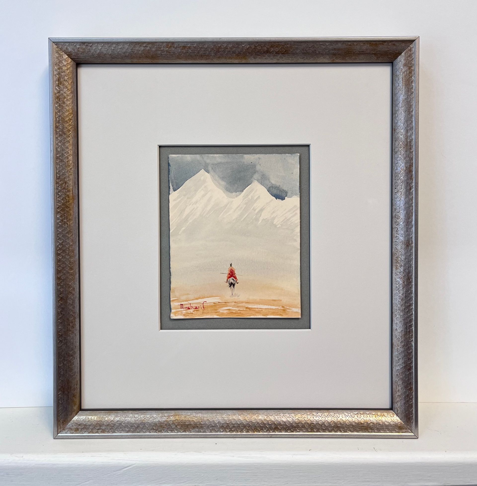 A small, framed watercolor painting shows a solitary figure in red walking across a vast, pale desert toward mountains.