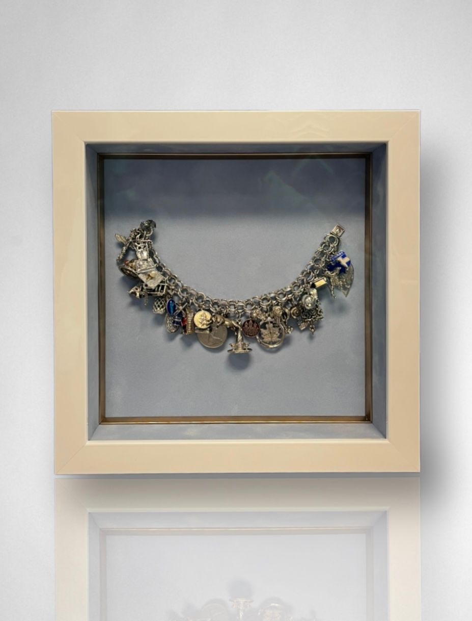 A silver charm bracelet with various trinkets displayed inside a square, cream-colored shadow box frame against a wall.