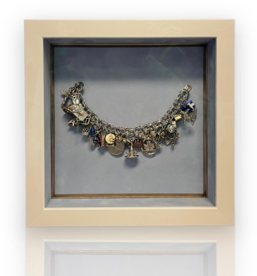 A vintage silver charm bracelet displayed inside a light-colored, square shadow box frame against a pale gray background.