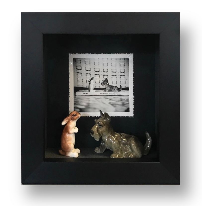 A black shadow box frame contains a vintage black-and-white photo of a dog above figurines of a rabbit and a terrier.