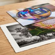 Three colorful abstract art prints stacked on a wooden table with a white vase in the background