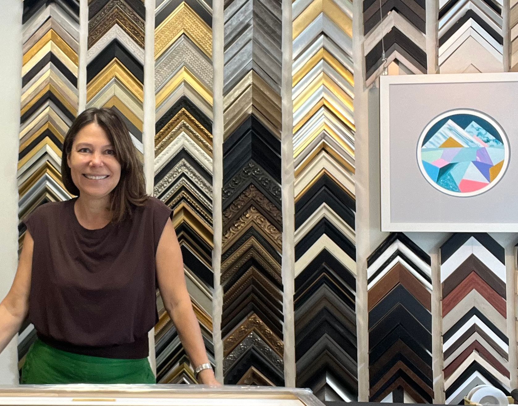 A person stands smiling in front of a wall covered in many different styles of picture frame samples.