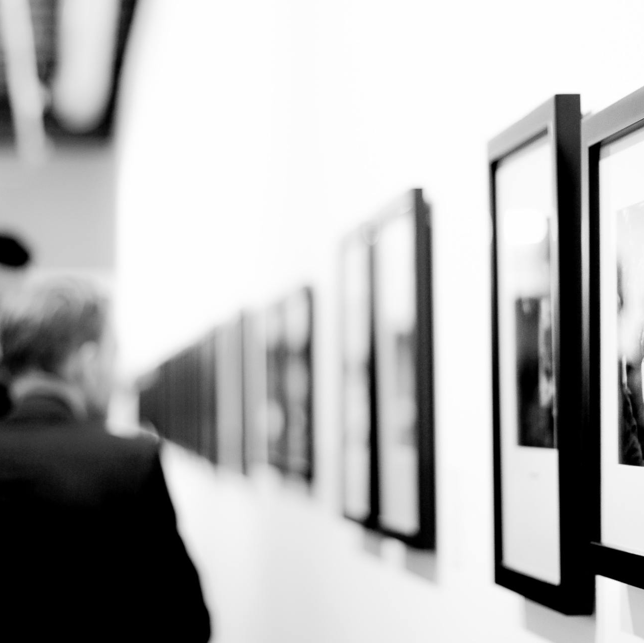 A person in a dark suit views a row of framed black-and-white photographs hanging on a white gallery wall.