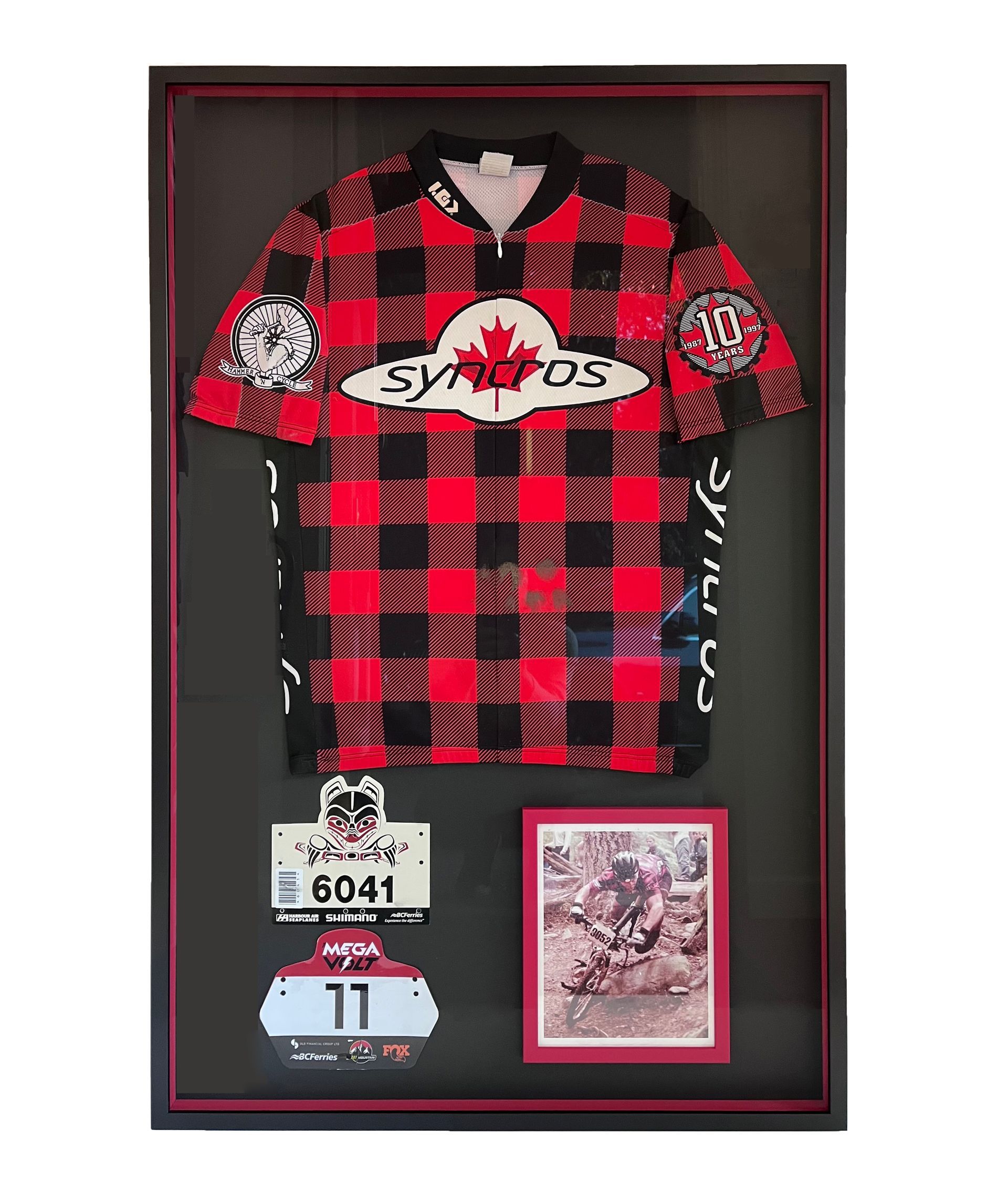 A framed display containing a red and black plaid Syncros cycling jersey, two race bibs, and a small photograph.