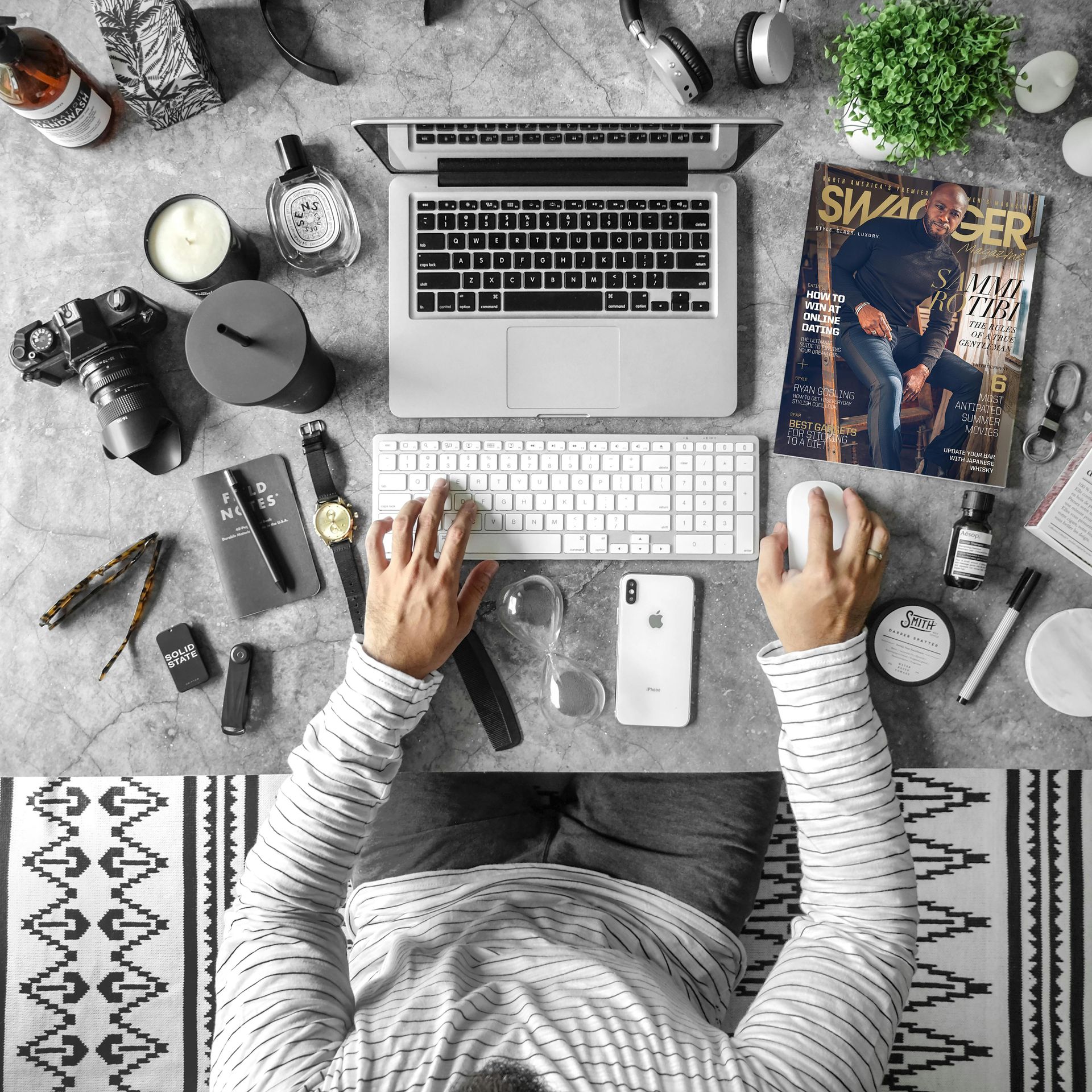 Overhead view of a person using a laptop and keyboard on a desk cluttered with office supplies, a camera, and a magazine.
