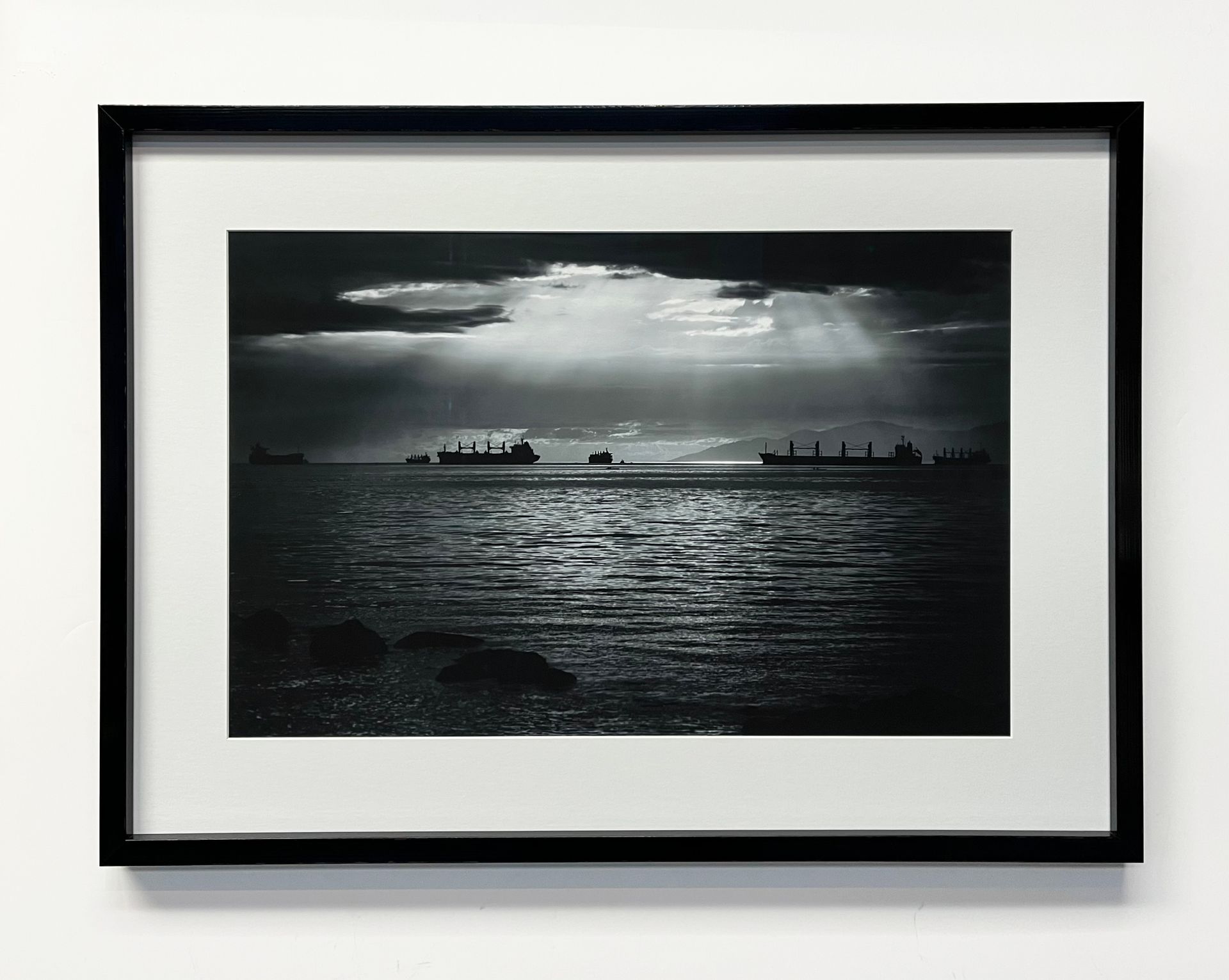 A framed, black-and-white landscape of ships on a dark, rippled sea under clouds with light rays shining down.