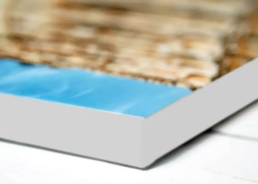 A close-up, angled view of a rectangular canvas print featuring a blue water surface meeting a textured, sandy shore.