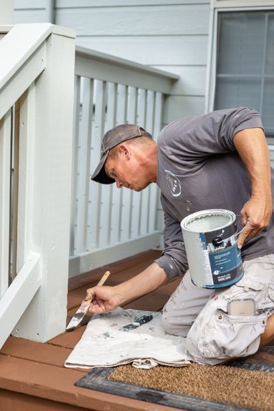 Man painting a white deck railing with a brush, kneeling outdoors.