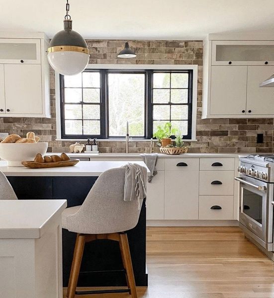 A modern kitchen with white cabinets, black trim windows, and a brick backsplash. A seated bar stool is in the foreground.