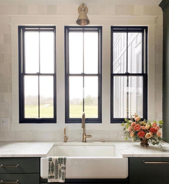 Kitchen sink under three windows, black trim, white countertop, green cabinets, gold faucet and hardware, flower arrangement.