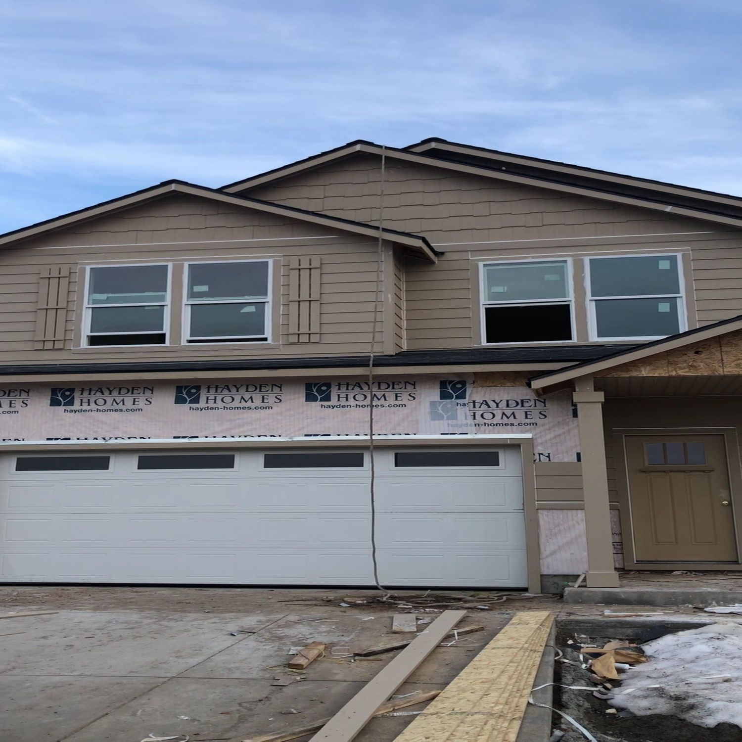 Two-story house under construction with beige siding, white garage doors, and snow on the ground