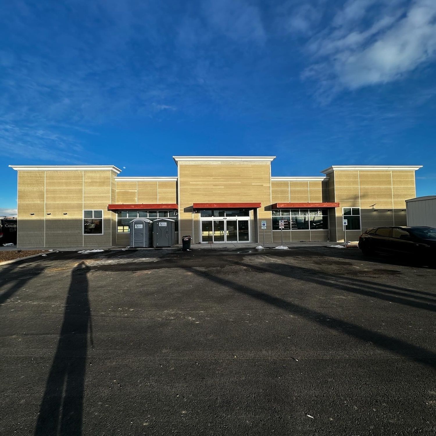 Empty storefront with beige facade and red awnings under a blue sky, viewed from a parking lot