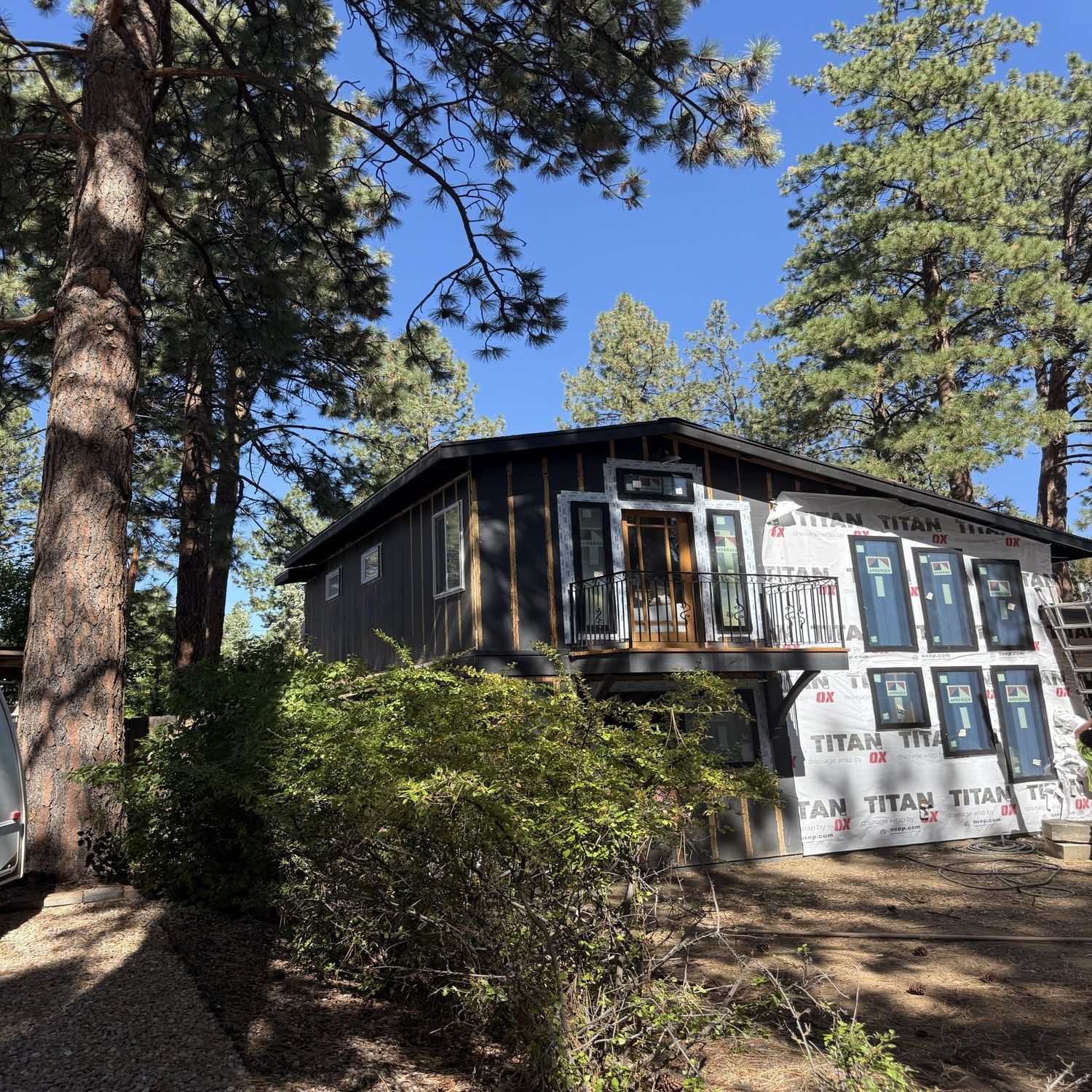 Forest cabin under construction with dark siding, white insulation panels, and tall pine trees around it