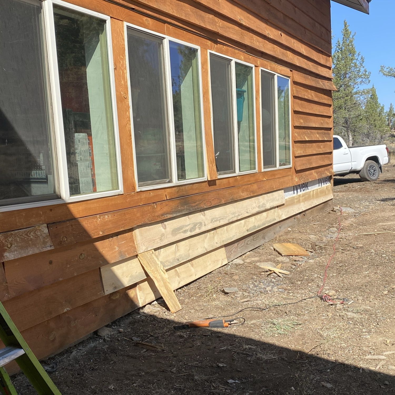 Wooden house exterior with large windows, a support brace, and a white pickup truck parked beside it.
