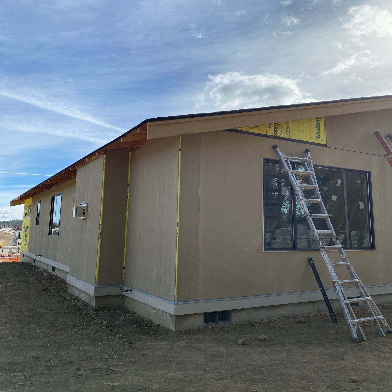 Single-story beige house under construction with ladder leaning against a window under a partly cloudy sky