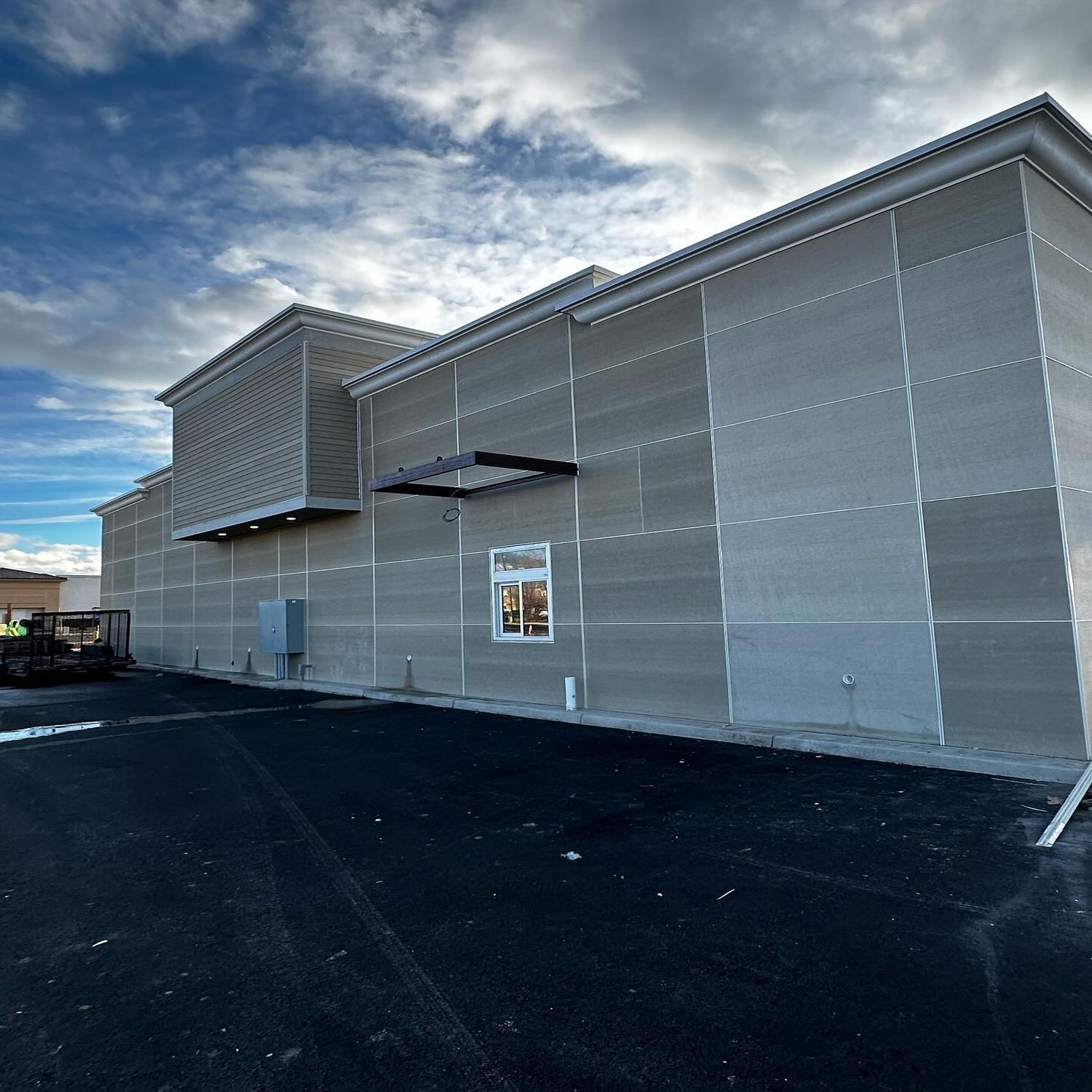 Large gray metal building exterior with a few windows and a poster near the entrance, beside a paved lot under a cloudy sky
