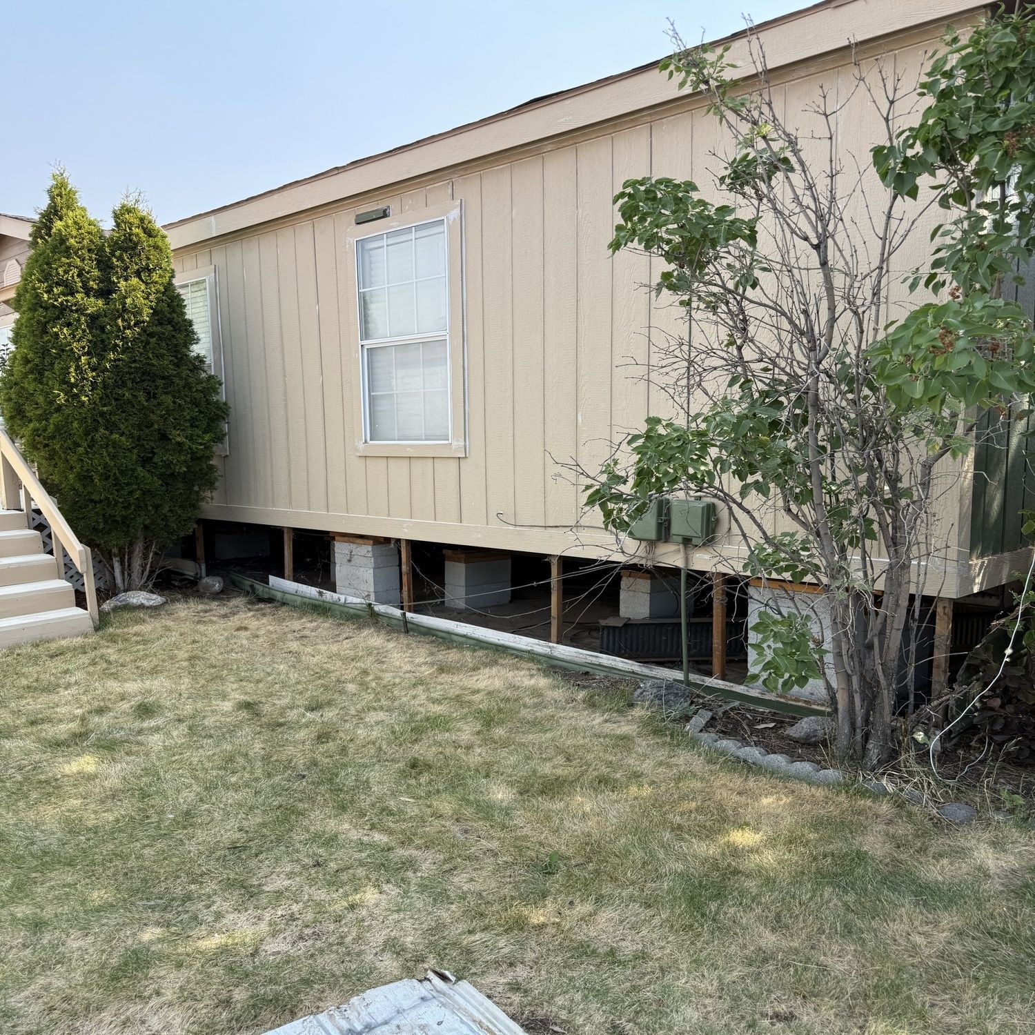 Beige mobile home on blocks with white window, trees, and dry grassy yard