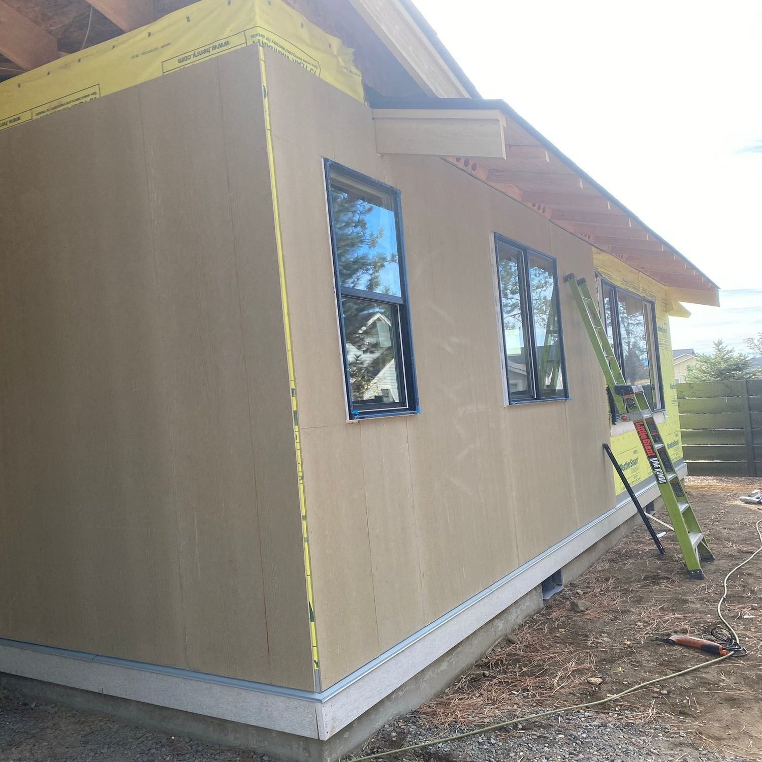 Small tan house exterior under construction with taped seams, windows, and a ladder leaning against the wall.