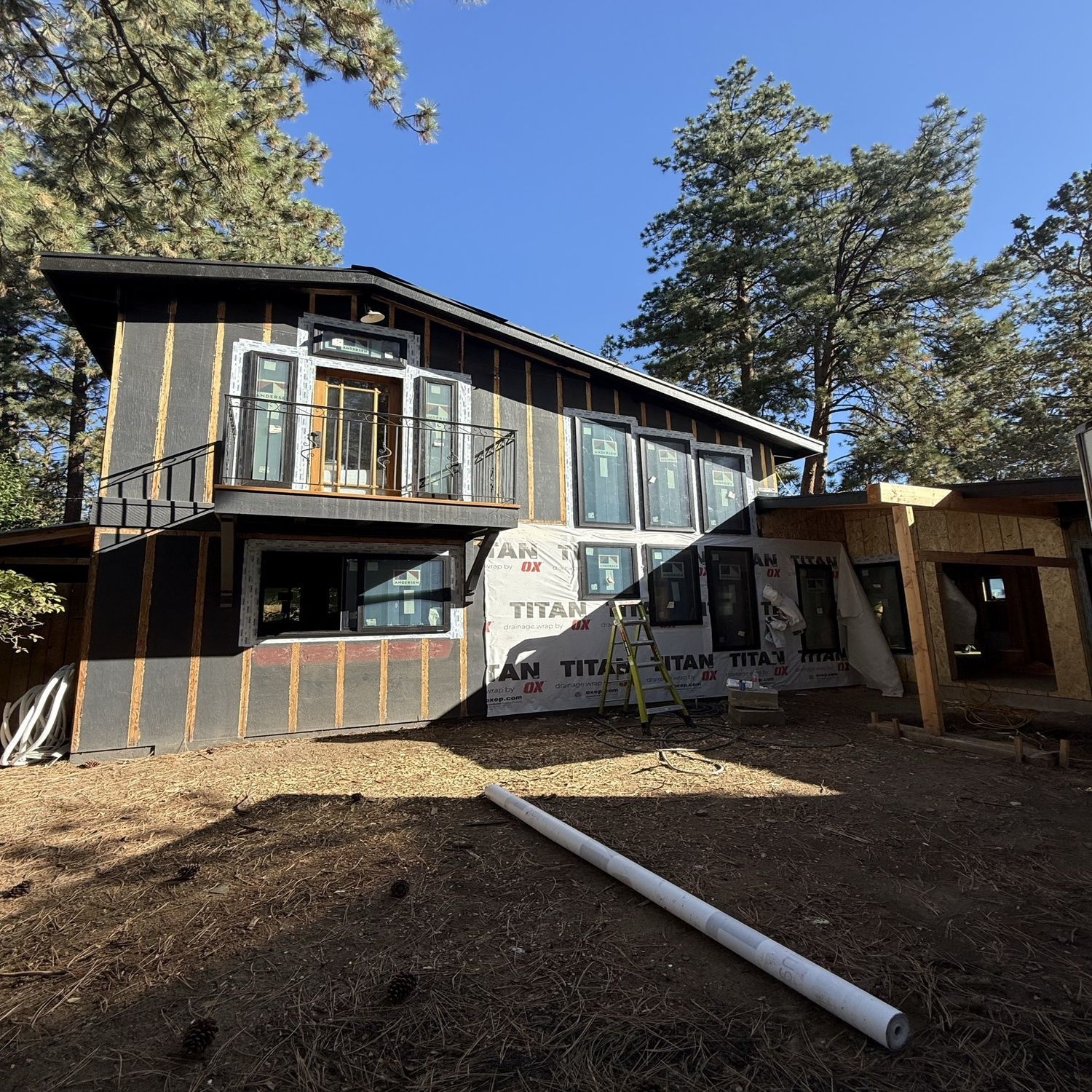 Modern two-story house under construction with exposed framing, wrap, and tall trees under a clear blue sky
